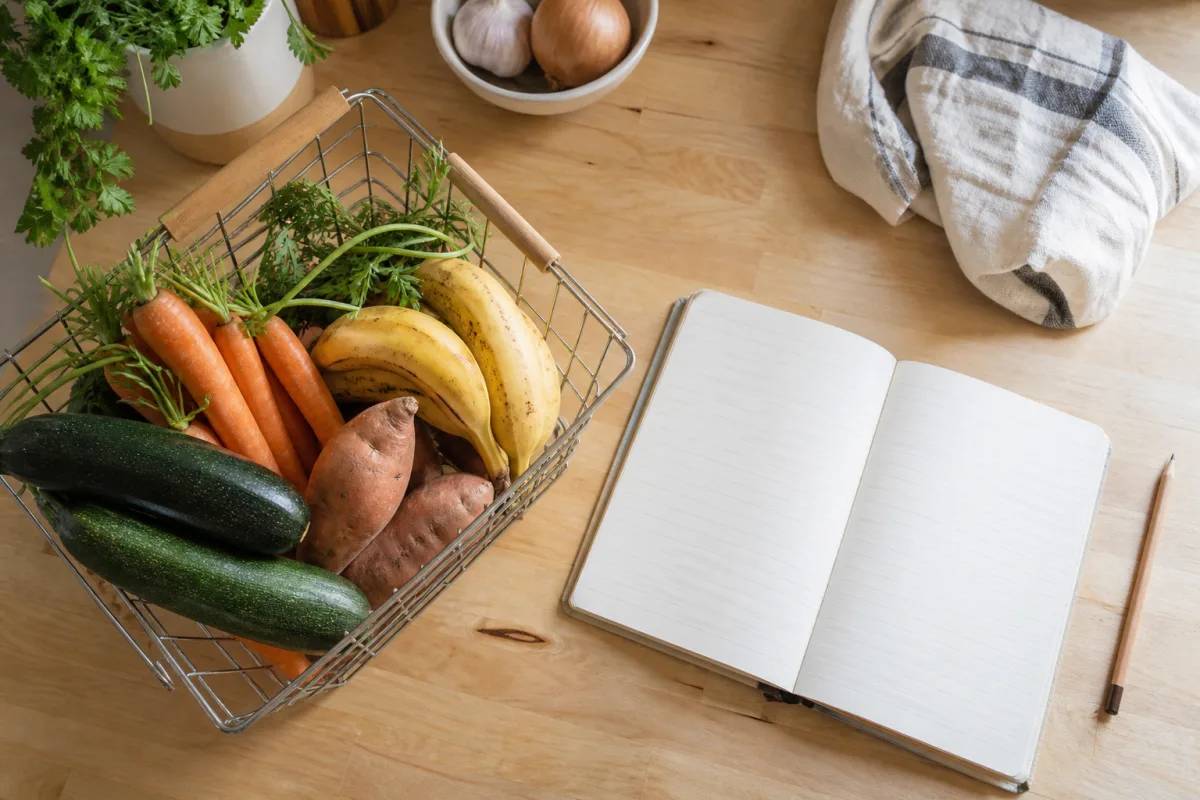 A notebook and pencil beside a basket of fresh produce on a kitchen counter