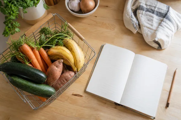 A notebook and pencil beside a basket of fresh produce on a kitchen counter