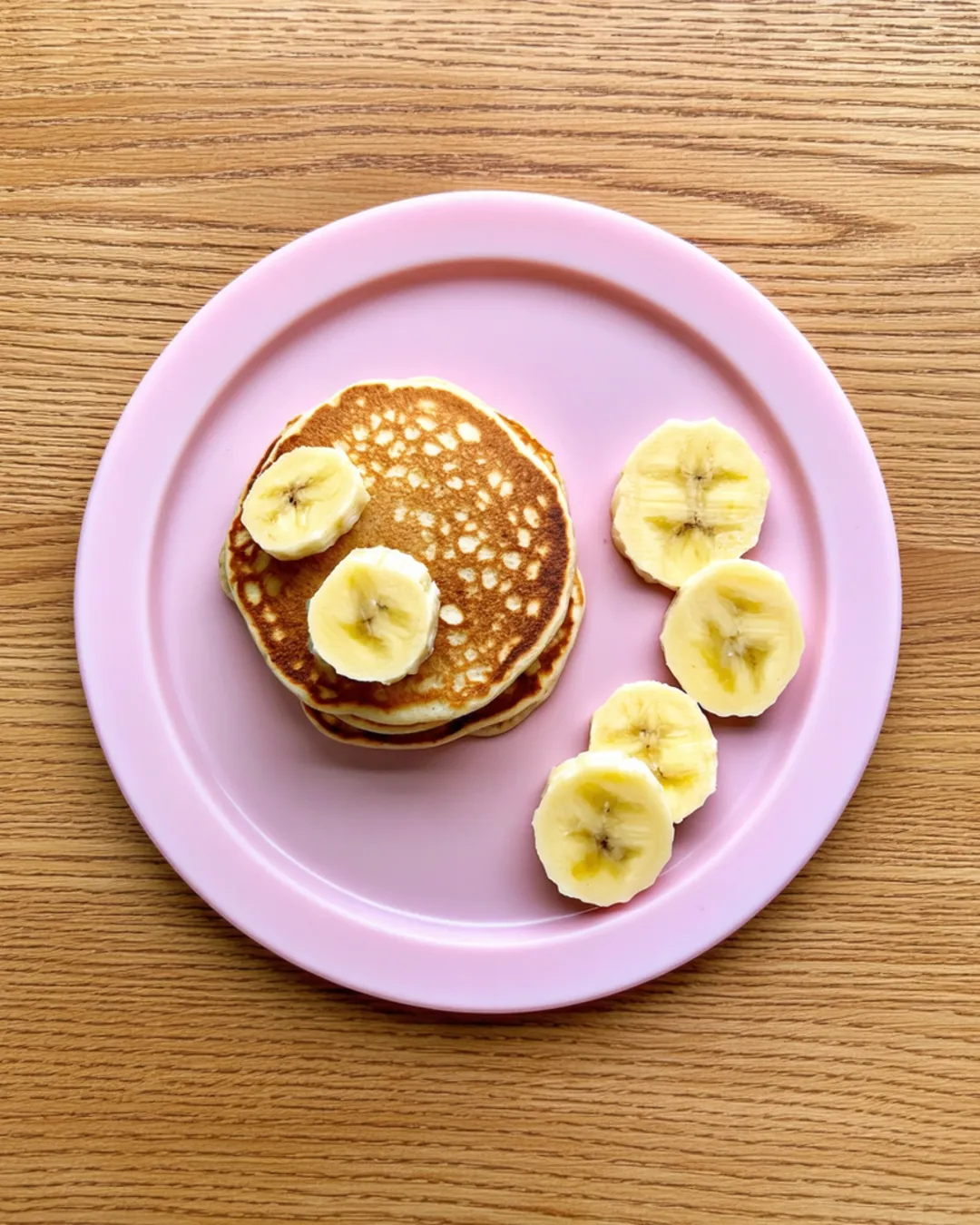 A small stack of mini banana oat pancakes with banana slices on a pink toddler plate
